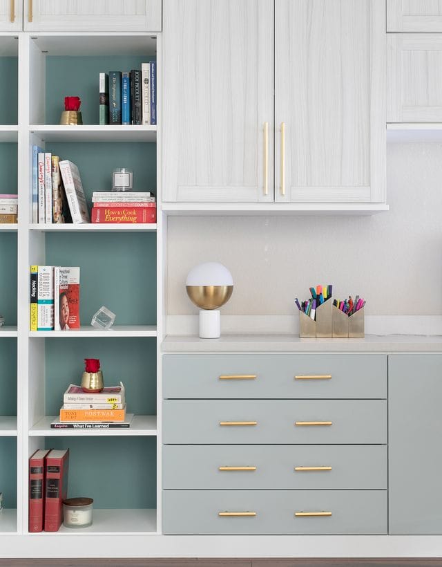 Custom bookshelves in light grey finish with back panels in blue and gold hardware handles by California Closets