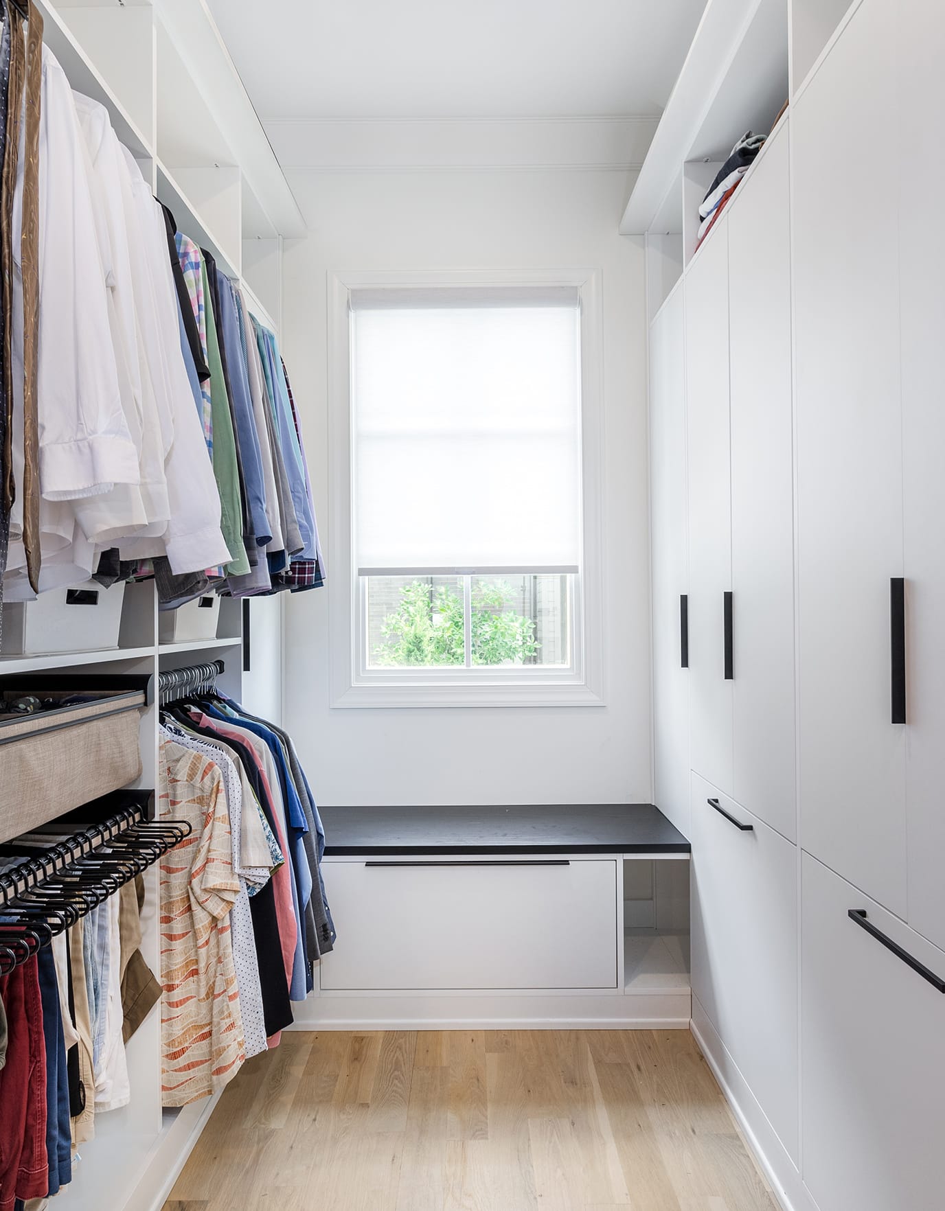 His custom walk in closet designed with extra storage and a window bench in white wood grain finish by California Closets