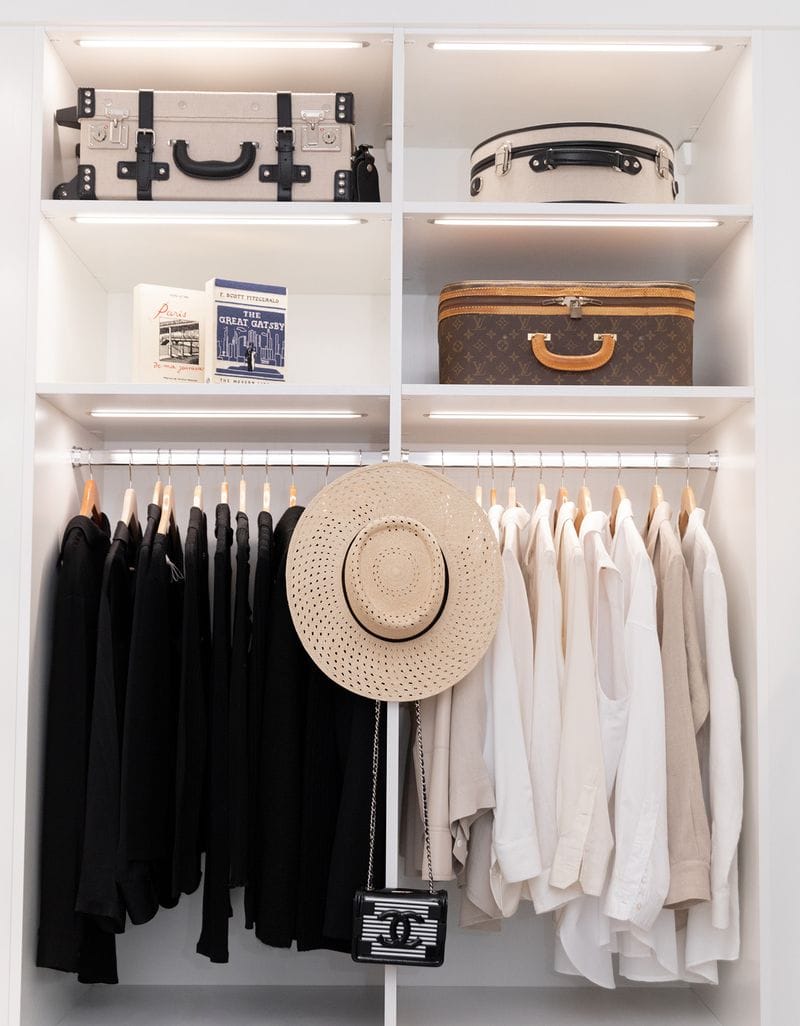 Closet shelving and hanging clothes poles under LED lighting in a white wood grain finish by California Closets